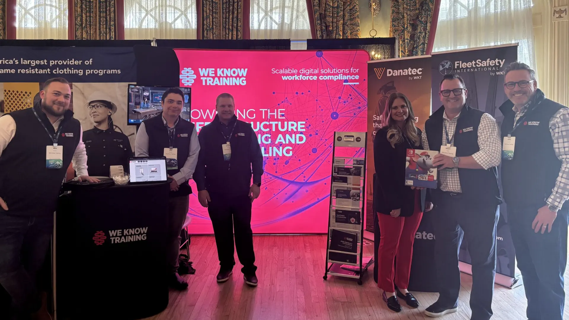 Six We Know Training team members at their pink trade-show booth, flanked by Danatec and Fleet Safety International banners.